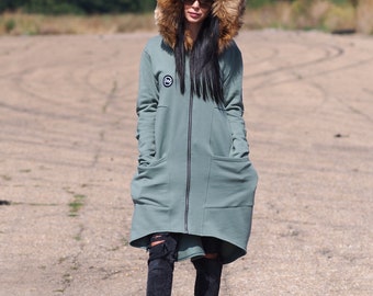 May include: A long, sage green hooded sweatshirt with a faux fur hood. The sweatshirt has a zipper closure and two large pockets. The model is wearing black jeans and black sneakers.