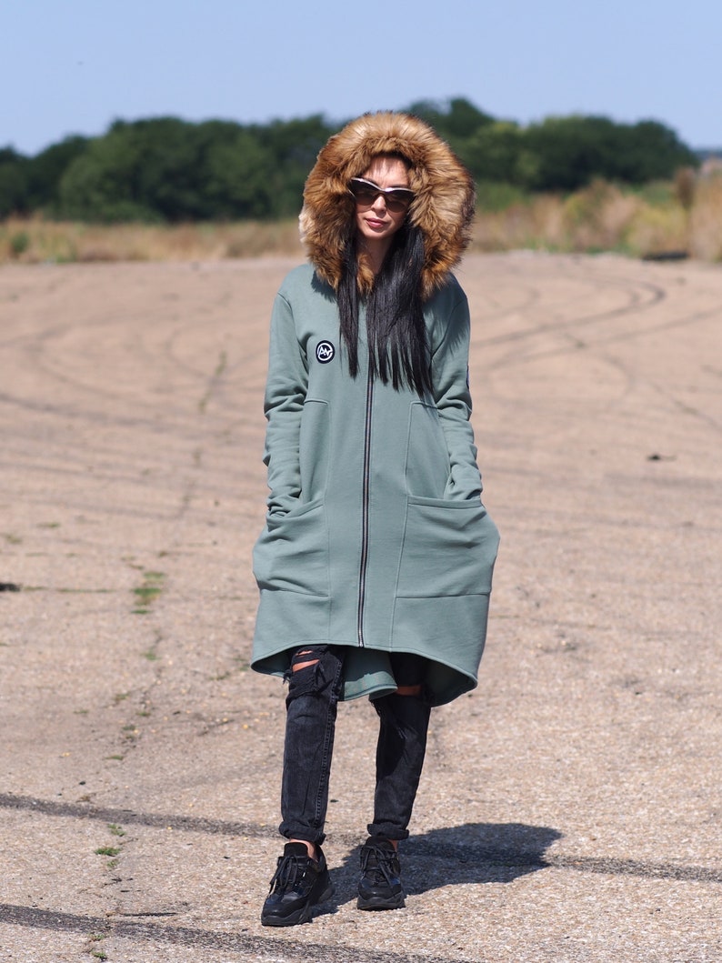 May include: A long, sage green hooded sweatshirt with a faux fur hood. The sweatshirt has a zipper closure and two large pockets. The model is wearing black jeans and black sneakers.