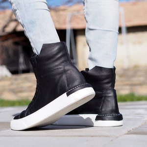 May include: A pair of black leather high-top sneakers with white soles. The sneakers are worn by a person wearing light blue jeans.