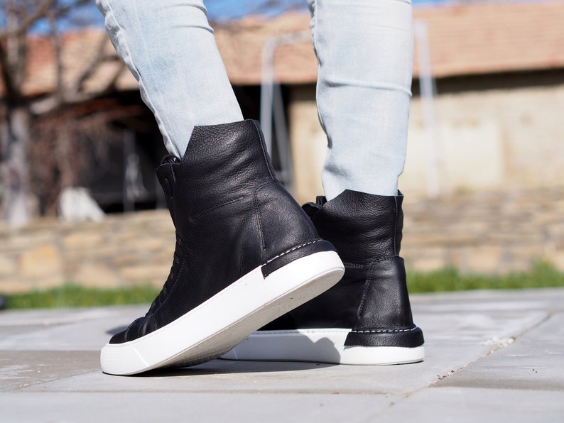 May include: A pair of black leather high-top sneakers with white soles. The sneakers are worn by a person wearing light blue jeans.