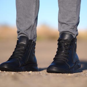 May include: Black leather high-top sneakers with laces and side zippers. The shoes have a thick, textured sole and are worn with gray denim jeans. The background is blurred, with a blue sky visible.