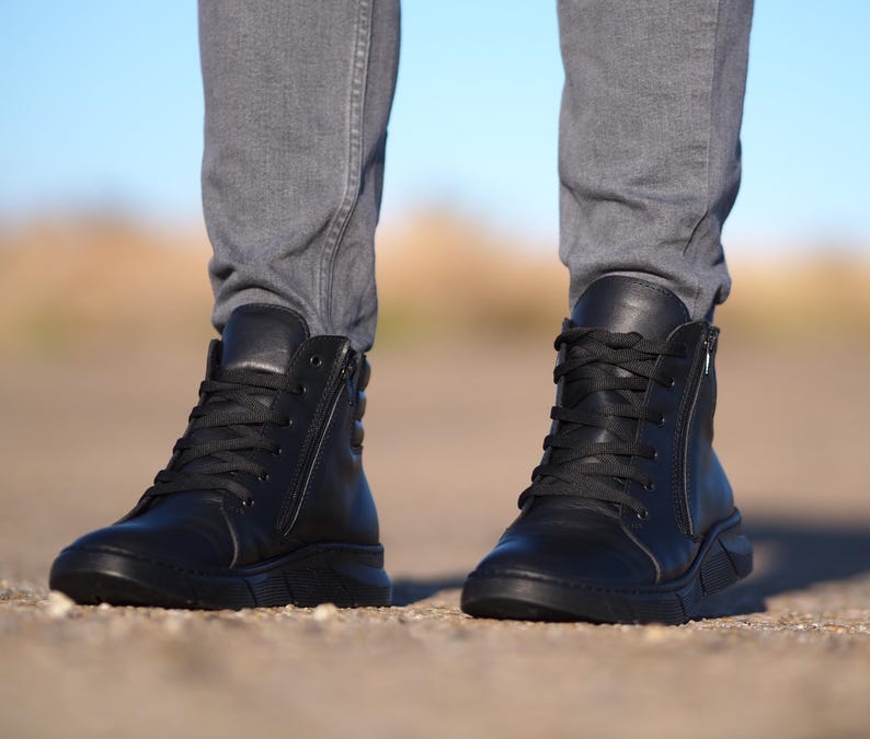 May include: Black leather high-top sneakers with laces and side zippers. The shoes have a thick, textured sole and are worn with gray denim jeans. The background is blurred, with a blue sky visible.