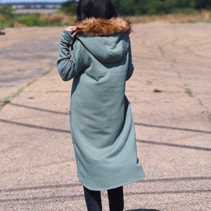 May include: A long, sage green hooded sweatshirt with a faux fur trim around the hood. The sweatshirt has a relaxed fit and a slightly curved hem.