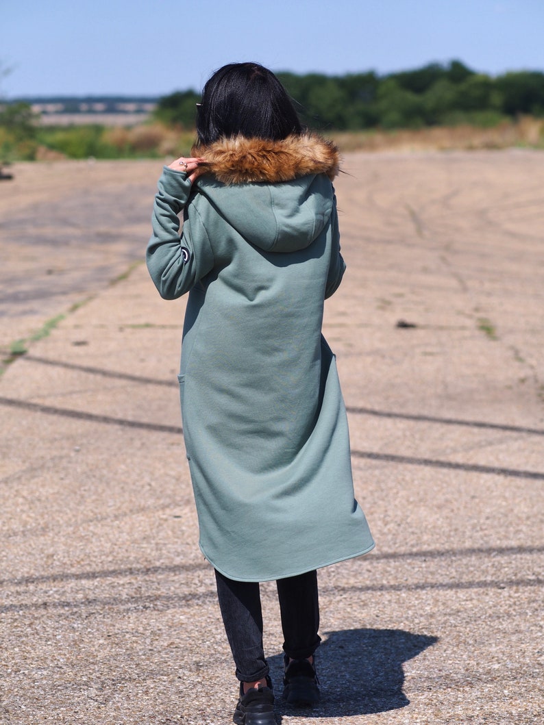May include: A long, sage green hooded sweatshirt with a faux fur trim around the hood. The sweatshirt has a relaxed fit and a slightly curved hem.
