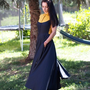 May include: A woman wearing a black maxi dress with a yellow and black collar. The dress has a flowy skirt and is cinched at the waist.