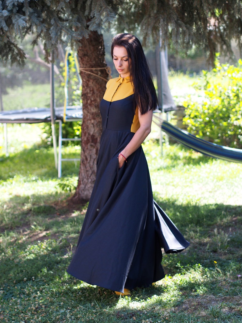 May include: A woman wearing a black maxi dress with a yellow and black collar. The dress has a flowy skirt and is cinched at the waist.