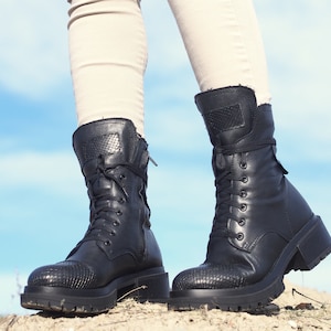 K&ouml;nnte beinhalten: Ein Paar schwarze Lederstiefel mit Schn&uuml;rung und einem dicken Absatz. Die Stiefel haben ein strukturiertes Muster an der Spitze und einen Rei&szlig;verschluss an der Seite.