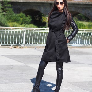 May include: A black long-sleeved coat with leather sleeves and a belt. The coat is worn over black leggings and black ankle boots.