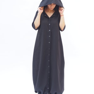 Puede incluir: Un vestido negro con capucha, con botones en la parte delantera y una falda larga y fluida. El vestido tiene un corte holgado y est&aacute; hecho de una tela suave y c&oacute;moda.