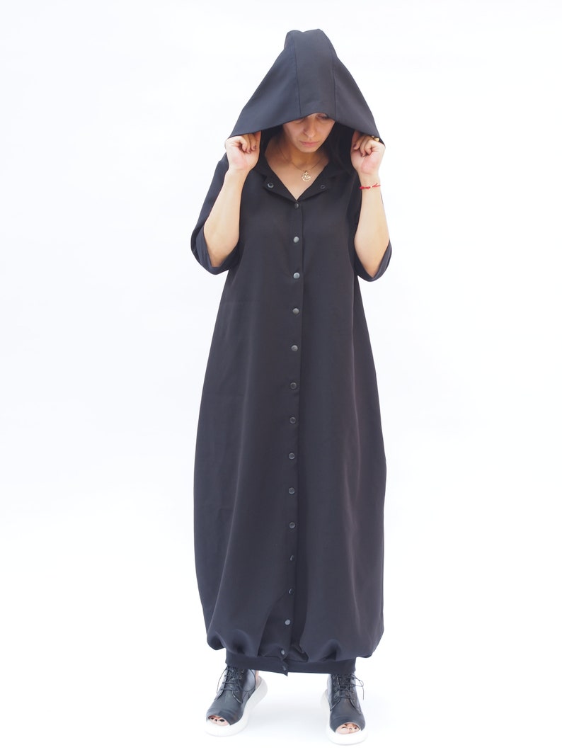 Puede incluir: Un vestido negro con capucha, con botones en la parte delantera y una falda larga y fluida. El vestido tiene un corte holgado y est&aacute; hecho de una tela suave y c&oacute;moda.