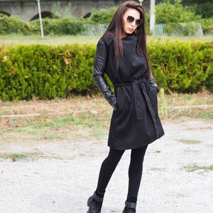 May include: A black long-sleeved coat with a leather trim on the sleeves. The coat has a belt and a collar that can be worn up or down. The model is wearing black leggings and black ankle boots.
