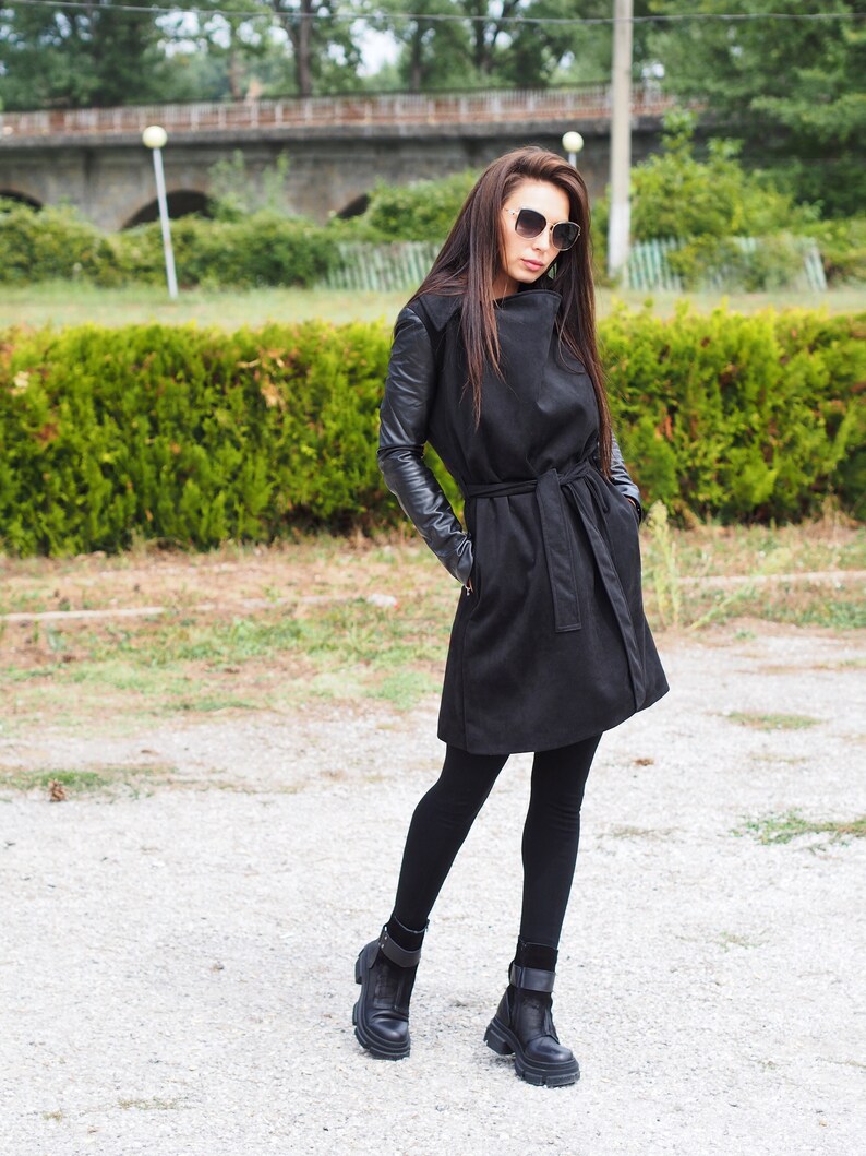 May include: A black long-sleeved coat with a leather trim on the sleeves. The coat has a belt and a collar that can be worn up or down. The model is wearing black leggings and black ankle boots.