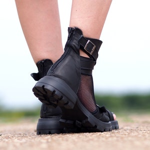 K&ouml;nnte beinhalten: Schwarze Ledersandalen mit Rei&szlig;verschluss an der R&uuml;ckseite und einem Schnallenriemen um den Kn&ouml;chel. Die Sandalen haben eine klobige Plateausohle und eine Netzsocke.