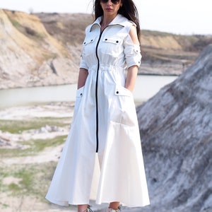 May include: A white long-sleeved dress with a black zipper down the front and a black drawstring at the waist. The dress has a relaxed fit and a flowing skirt. The model is wearing the dress with white sneakers.
