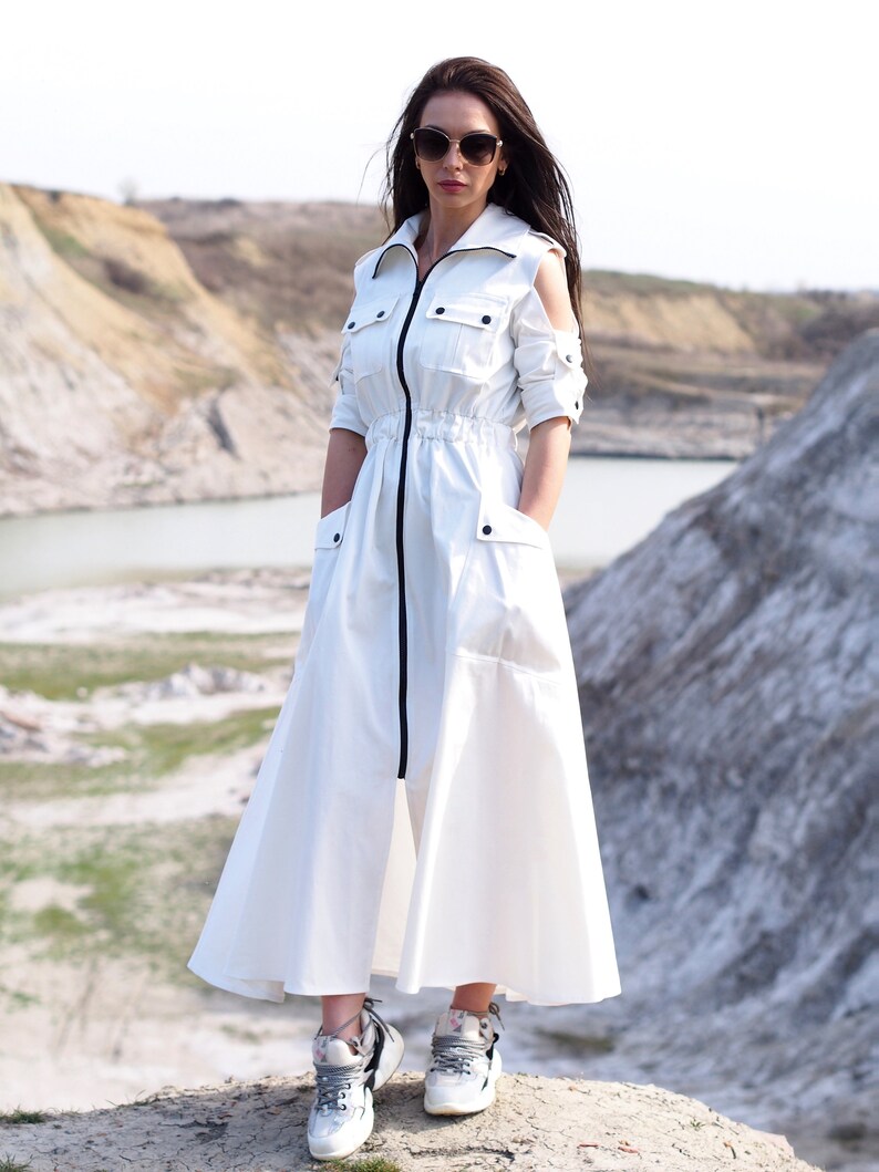 May include: A white long-sleeved dress with a black zipper down the front and a black drawstring at the waist. The dress has a relaxed fit and a flowing skirt. The model is wearing the dress with white sneakers.
