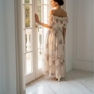 May include: A cream-colored off-the-shoulder maxi dress with a floral pattern. The dress features ruffled detailing and a tiered skirt. The woman is standing near a white door with a gold handle, and the floor is marble.