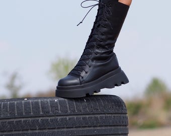 May include: Black leather lace-up boots with a chunky platform sole. The boots are being worn by a person standing on a black tyre.