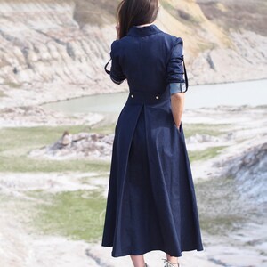 May include: A woman wearing a long, dark blue denim dress with a button-up collar and long sleeves. She is standing on a rocky hilltop with a body of water in the background.