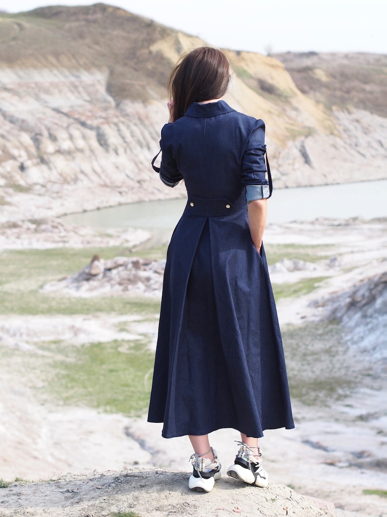 May include: A woman wearing a long, dark blue denim dress with a button-up collar and long sleeves. She is standing on a rocky hilltop with a body of water in the background.