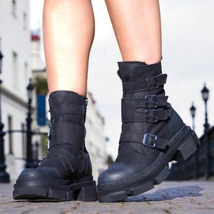 May include: Black ankle boots with multiple straps and buckles. The boots have a chunky sole and a zipper on the side. The boots are made of a matte material and are worn outdoors.