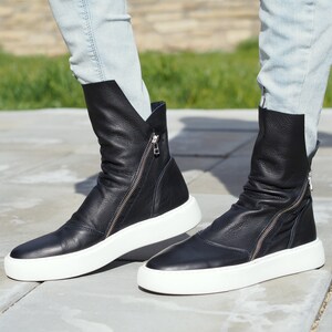 Puede incluir: Un par de botines negros de cuero con suelas blancas y cremalleras laterales. Las botas las lleva una persona que lleva unos vaqueros azules claros.