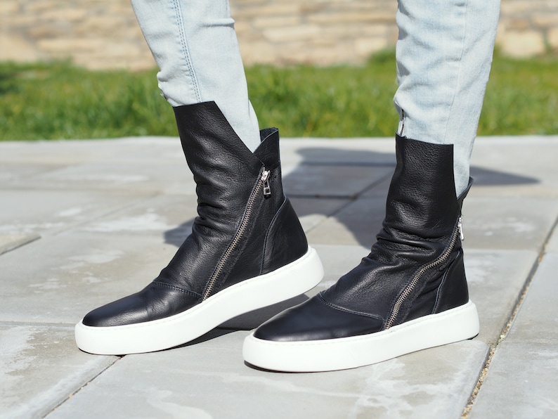 Puede incluir: Un par de botines negros de cuero con suelas blancas y cremalleras laterales. Las botas las lleva una persona que lleva unos vaqueros azules claros.