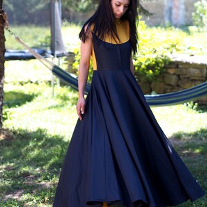 May include: A woman wearing a black and yellow dress with a full skirt. She is standing in a grassy area with a hammock in the background.