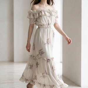 May include: An off-the-shoulder maxi dress in a light cream color featuring a delicate purple floral print. The garment includes a ruffled neckline, a matching fabric belt with a rectangular buckle at the waist, and a ruffled hemline.