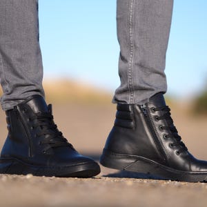 May include: Black leather high-top boots with laces and side zippers. The boots have a padded ankle and a thick sole. The boots are worn with gray jeans. The background is blurred with a blue sky.