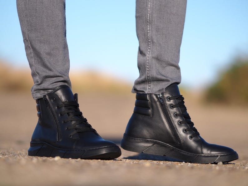 May include: Black leather high-top boots with laces and side zippers. The boots have a padded ankle and a thick sole. The boots are worn with gray jeans. The background is blurred with a blue sky.