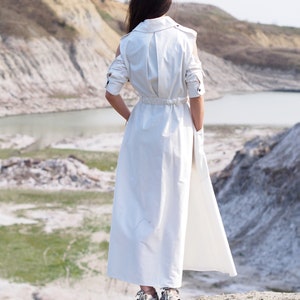 May include: A woman wearing a white long-sleeved dress with a cinched waist and a flowing skirt stands on a rocky hill overlooking a body of water.