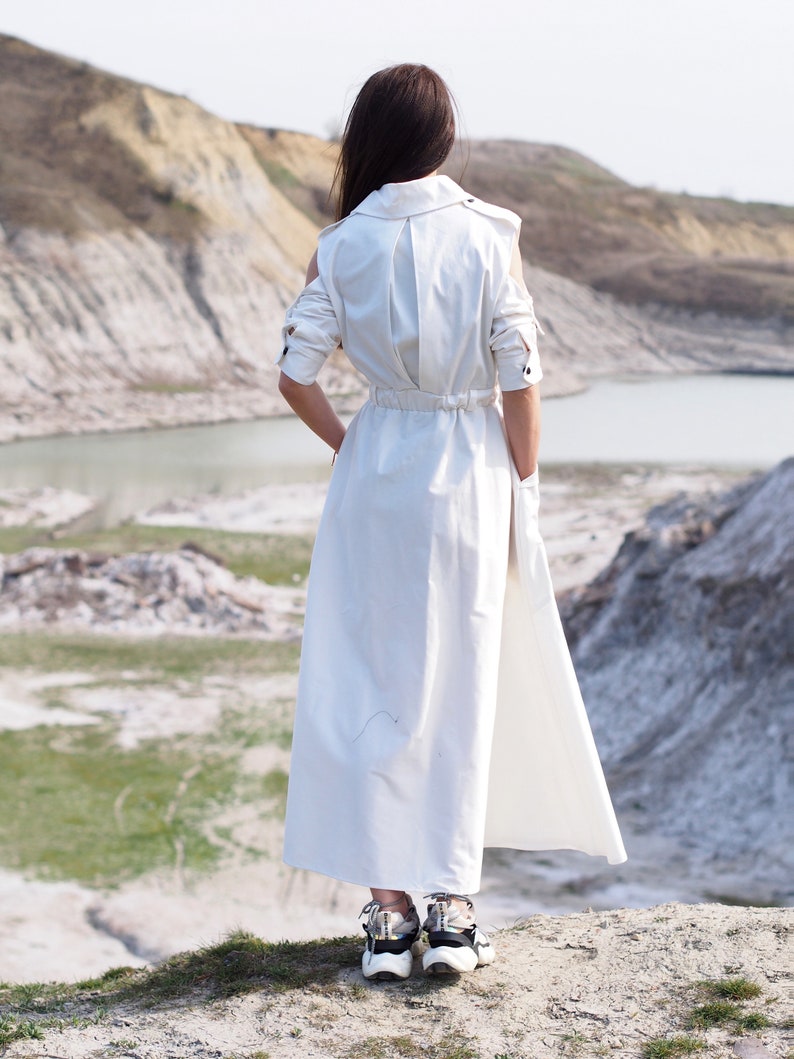 May include: A woman wearing a white long-sleeved dress with a cinched waist and a flowing skirt stands on a rocky hill overlooking a body of water.