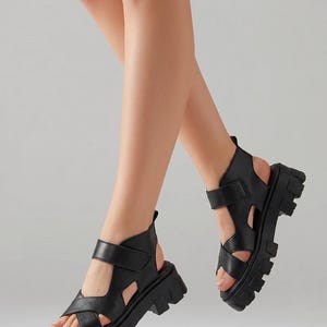 Pu&ograve; includere: Sandali neri con plateau, suola spessa e cinturini regolabili. I sandali hanno un design a punta aperta e un supporto alto alla caviglia. Le scarpe sono realizzate in materiale liscio e sono mostrate su uno sfondo grigio chiaro.