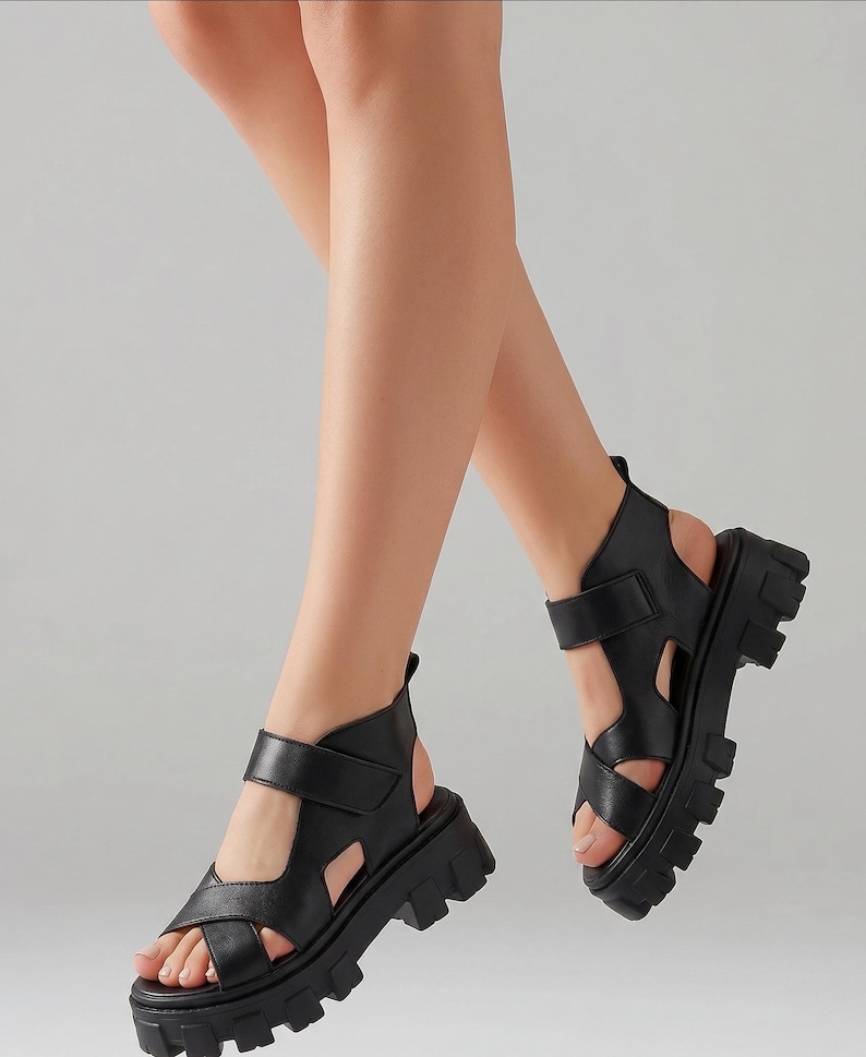Pu&ograve; includere: Sandali neri con plateau, suola spessa e cinturini regolabili. I sandali hanno un design a punta aperta e un supporto alto alla caviglia. Le scarpe sono realizzate in materiale liscio e sono mostrate su uno sfondo grigio chiaro.