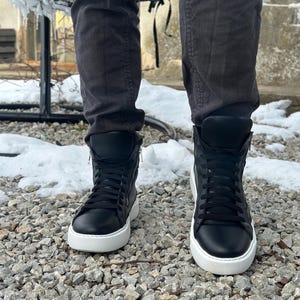 May include: Black high-top trainers with white soles and laces. The trainers have a zip on the side and are being worn by someone standing on a gravel surface.