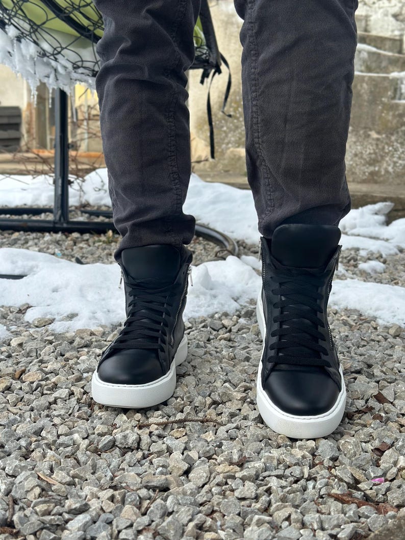 May include: Black high-top trainers with white soles and laces. The trainers have a zip on the side and are being worn by someone standing on a gravel surface.