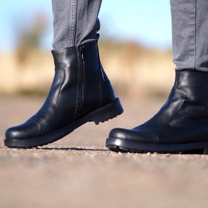 May include: A pair of black leather ankle boots with a side zipper. The boots have a rounded toe and a thick, textured sole. The person is wearing gray pants.