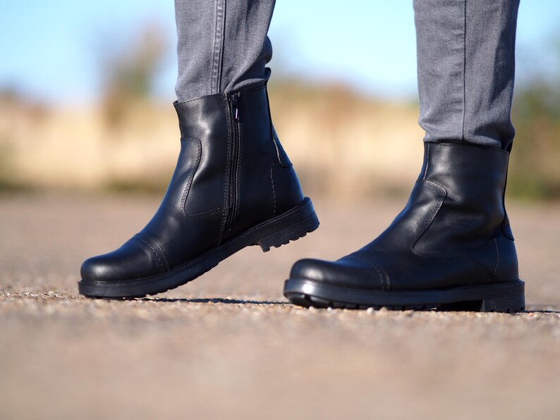 May include: A pair of black leather ankle boots with a side zipper. The boots have a rounded toe and a thick, textured sole. The person is wearing gray pants.