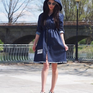 May include: A navy blue denim dress with a hood and a drawstring waist. The dress has white text around the hem that reads "Adventure Awaits You, Your Vibe Attracts Your Tribe." The model is wearing black sneakers.