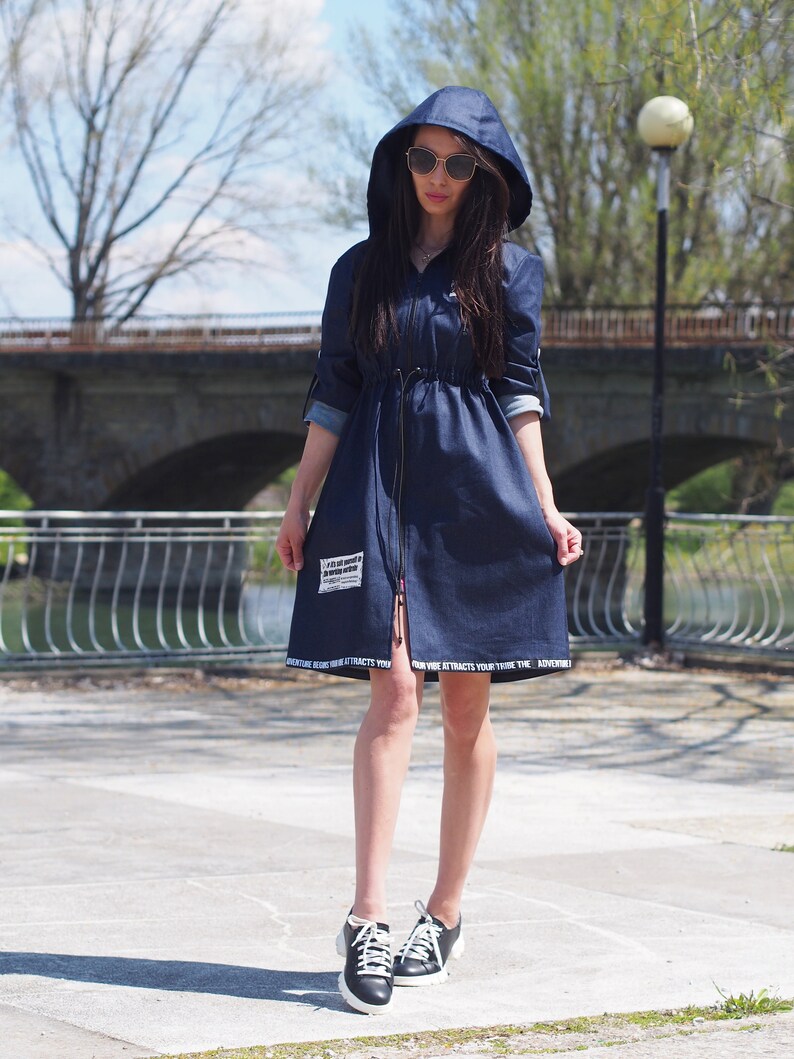 May include: A navy blue denim dress with a hood and a drawstring waist. The dress has white text around the hem that reads "Adventure Awaits You, Your Vibe Attracts Your Tribe." The model is wearing black sneakers.