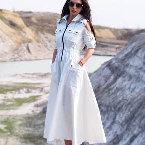 May include: A white long-sleeved dress with a zipper closure, a drawstring waist, and two pockets. The dress has a relaxed fit and a flowing skirt. The model is wearing sunglasses and grey sneakers.
