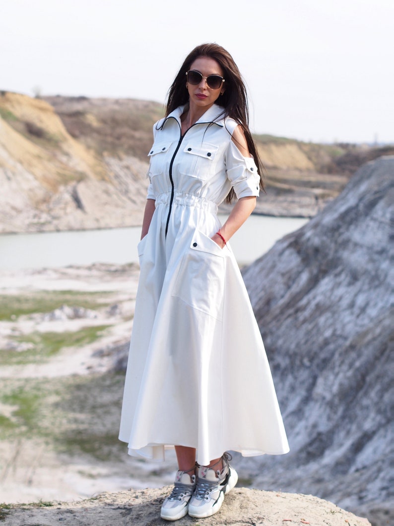 May include: A white long-sleeved dress with a zipper closure, a drawstring waist, and two pockets. The dress has a relaxed fit and a flowing skirt. The model is wearing sunglasses and grey sneakers.