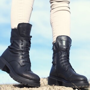 K&ouml;nnte beinhalten: Ein Paar schwarze Lederstiefel mit Schn&uuml;rsenkeln und einem strukturierten Muster an der Spitze. Die Stiefel haben einen dicken Absatz und werden von einer Person getragen, die auf einem Feldweg steht.