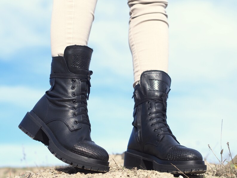 K&ouml;nnte beinhalten: Ein Paar schwarze Lederstiefel mit Schn&uuml;rsenkeln und einem strukturierten Muster an der Spitze. Die Stiefel haben einen dicken Absatz und werden von einer Person getragen, die auf einem Feldweg steht.