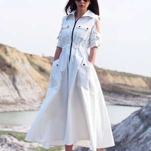 May include: A white long-sleeved dress with a zipper closure, a drawstring waist, and two large pockets. The dress has cut-out shoulders and a flowing skirt. The model is wearing the dress with white sneakers.