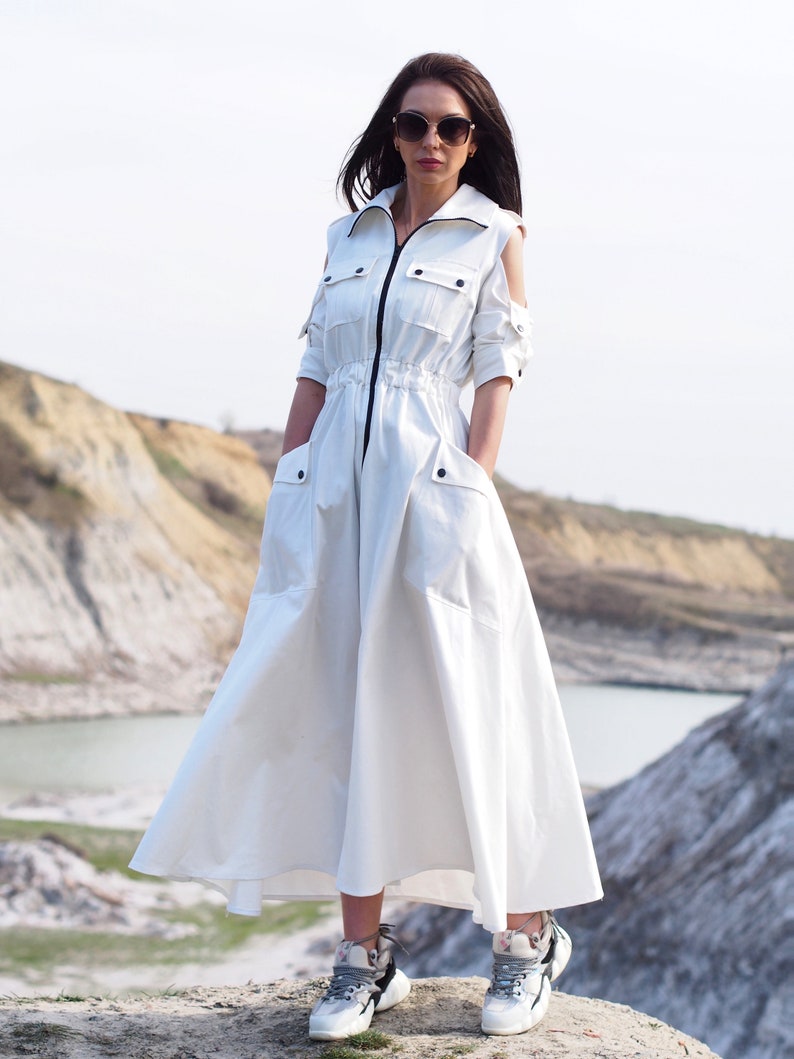 May include: A white long-sleeved dress with a zipper closure, a drawstring waist, and two large pockets. The dress has cut-out shoulders and a flowing skirt. The model is wearing the dress with white sneakers.