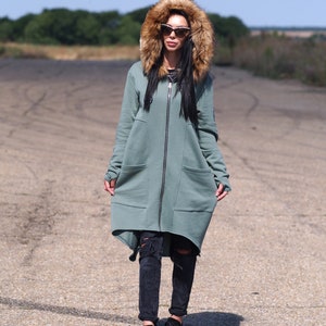 May include: A long, sage green hooded sweatshirt with a faux fur hood. The sweatshirt has a zipper closure and two large pockets. The model is wearing black jeans and black sneakers.