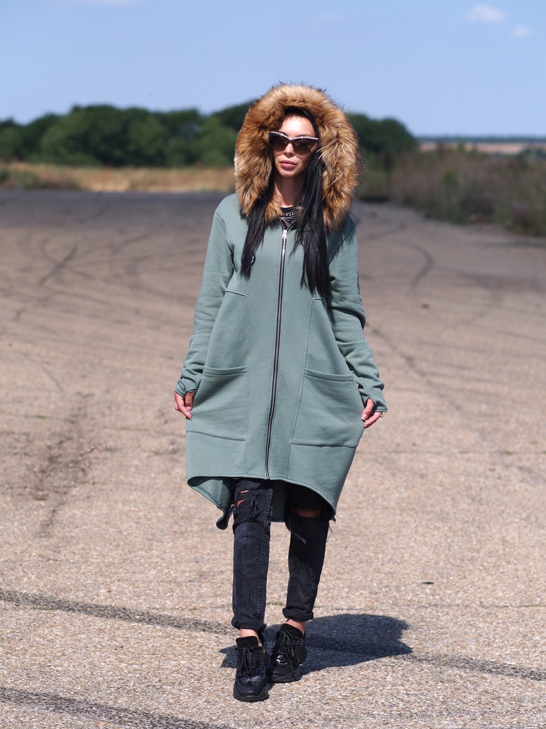 May include: A long, sage green hooded sweatshirt with a faux fur hood. The sweatshirt has a zipper closure and two large pockets. The model is wearing black jeans and black sneakers.