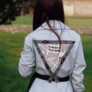 May include: A light blue long-sleeved shirt with a black belt and a newspaper print design on the back. The print includes the text "The Fashion Trend Fashion" and "Ry Mackay on "Great fightba".