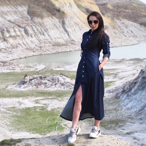 May include: A woman wearing a dark blue denim dress with a long slit up the side and white sneakers. She is standing on a rocky hill with a body of water in the background.